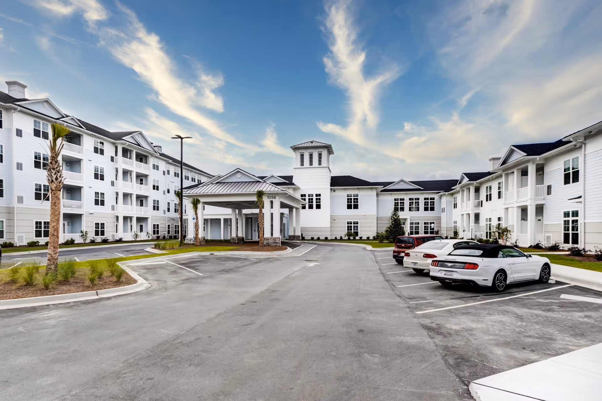 Exterior view of a senior living facility named Magnolia Bridge at Murrells Inlet, showing a large white multi-story building with balconies, a covered entrance, palm trees, a parking lot with several cars, and a partly cloudy blue sky.