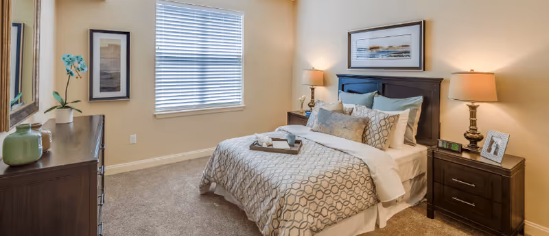 Well-lit furnished bedroom with a bed, bedside tables and lamps, a dresser and a window.