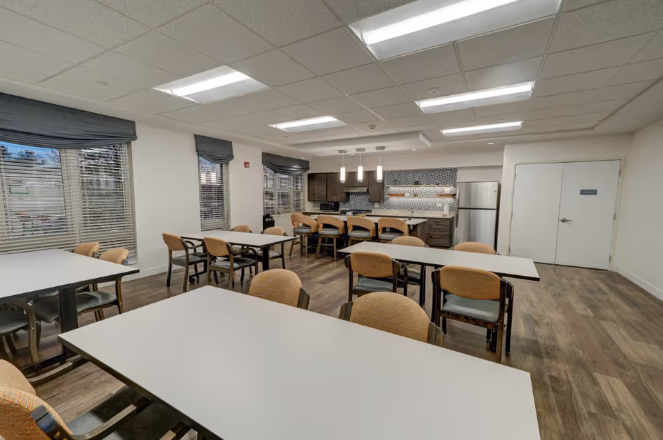 A spacious dining area in a senior living facility with multiple tables and chairs arranged neatly. The room features large windows with blinds and valances, wood flooring, and a kitchen area in the back with a refrigerator, cabinets, and a patterned backsplash. Pendant lights hang over a counter with bar stools.