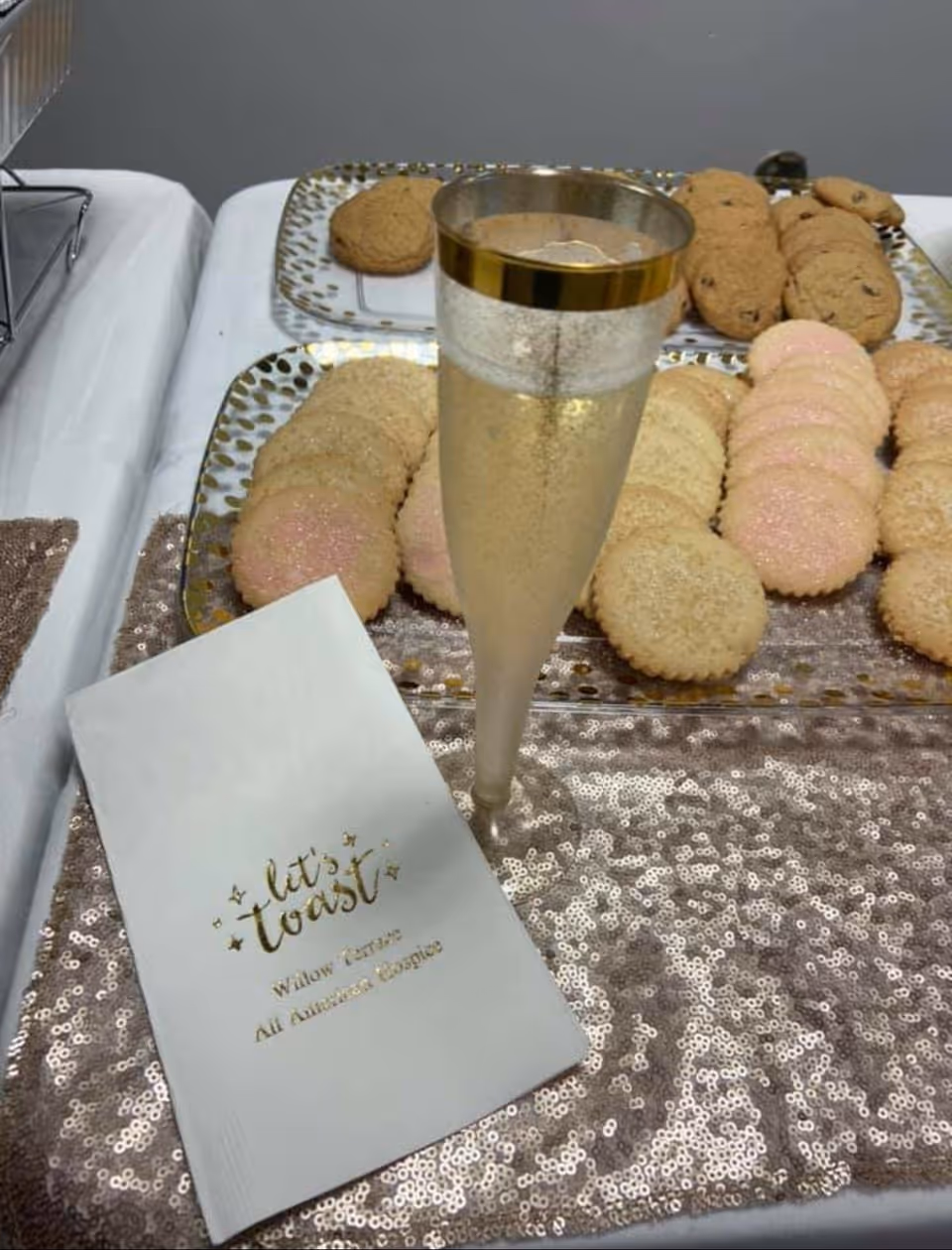 A close-up of a table setting with a glass of champagne, trays of assorted cookies, and a white napkin with gold text that reads 'let's toast Willow Terrace All Assisted Lifestyles' on a sparkly sequin table runner.