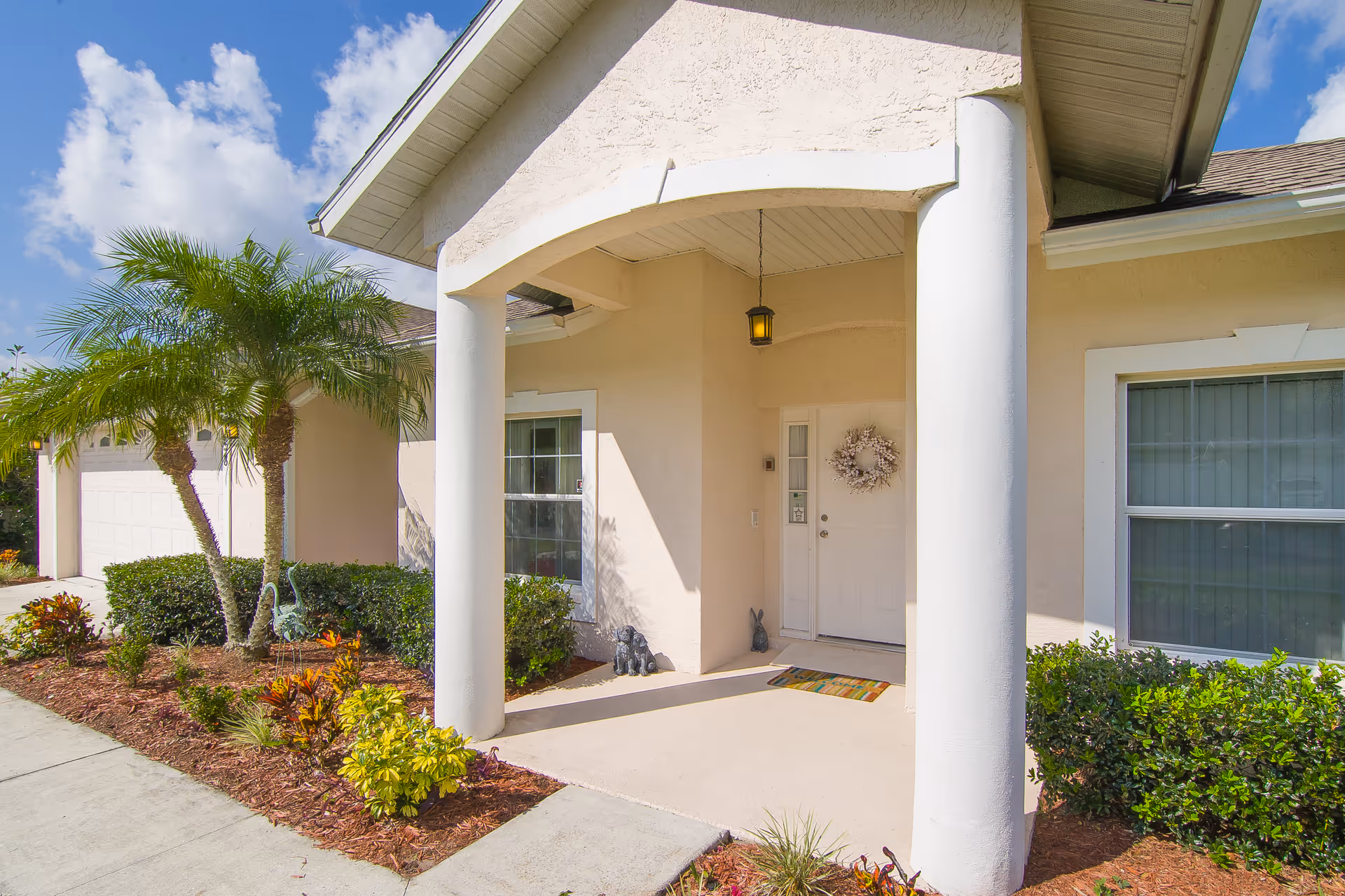 Front entrance of a beige-colored house with a covered porch supported by two white columns, a white door decorated with a wreath, a doormat, and small decorative statues. There are two windows on either side of the door, a hanging lantern light, and landscaping with palm trees and shrubs along the walkway.