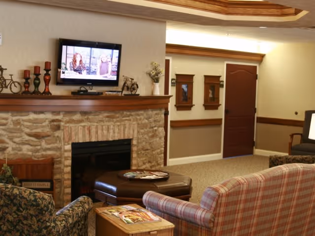 Cozy common living room with a stone fireplace, wall-mounted TV, and upholstered chairs and ottoman.