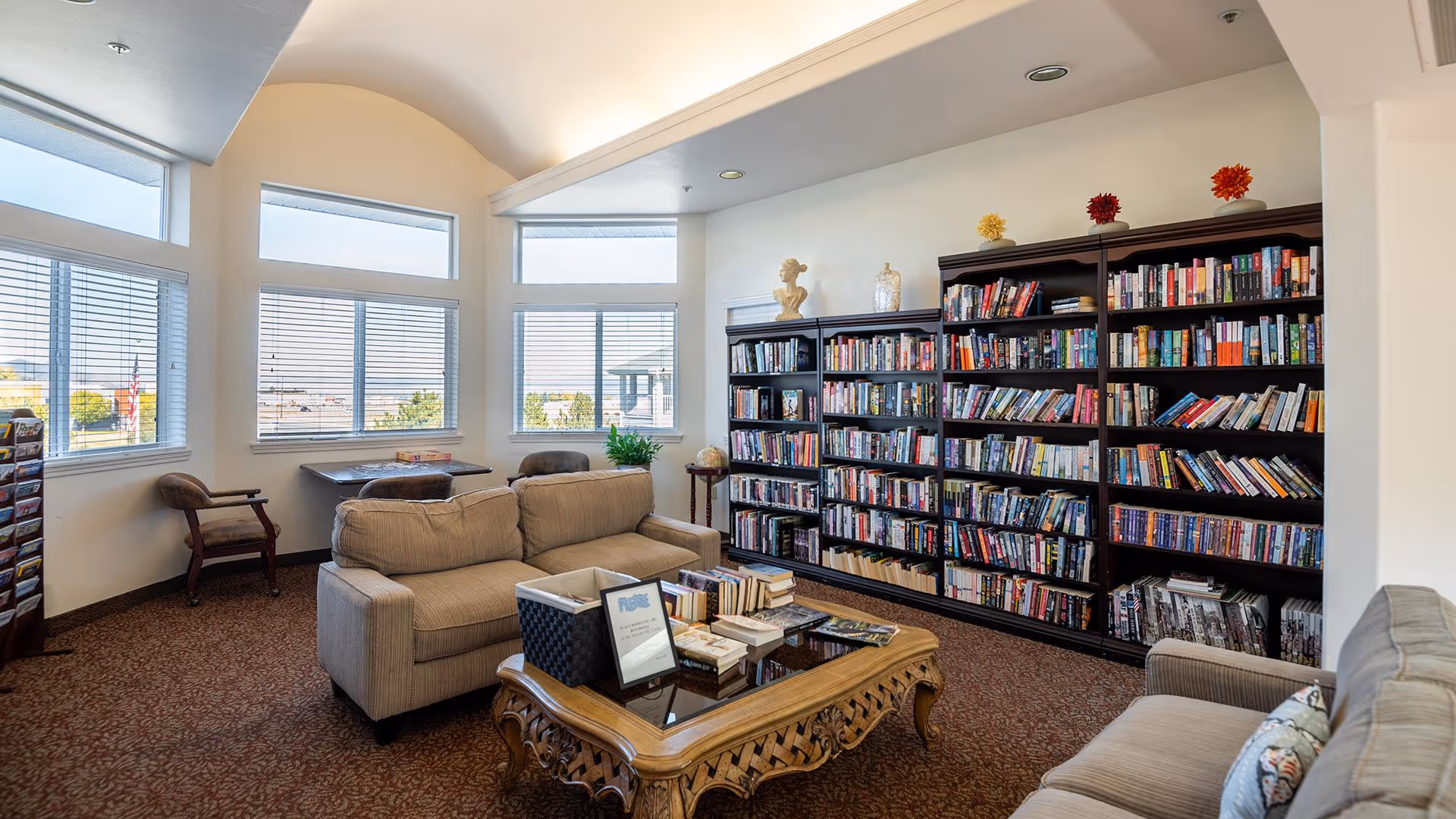 A cozy senior living facility library and lounge area with large windows letting in natural light. The room features two beige sofas facing a wooden coffee table with books and a basket on it. A large bookshelf filled with books lines one wall, and there are a couple of chairs and a small table near the windows.