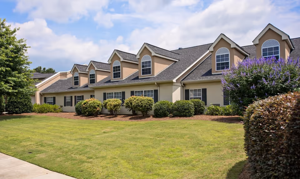 Exterior view of a beige senior living facility building with multiple dormer windows, surrounded by neatly trimmed bushes and a well-maintained green lawn under a partly cloudy sky.