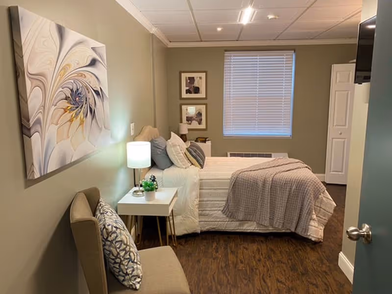 A cozy bedroom with a neatly made bed featuring white and gray bedding, a bedside table with a lamp and small plant, a cushioned chair with a patterned pillow, abstract wall art, two framed pictures on the wall, a window with blinds, and a wooden floor.