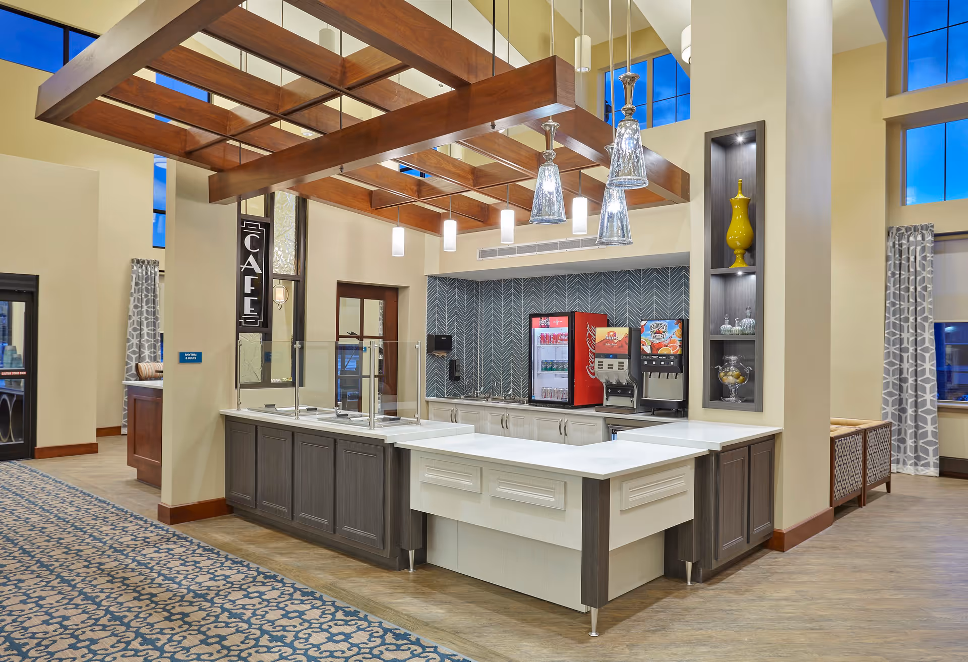 Interior view of a modern cafe area in a senior living facility with a serving counter, beverage dispensers including a Coca-Cola fridge and a juice machine, pendant lights hanging from a wooden beam ceiling, patterned carpet, and decorative shelving with vases.