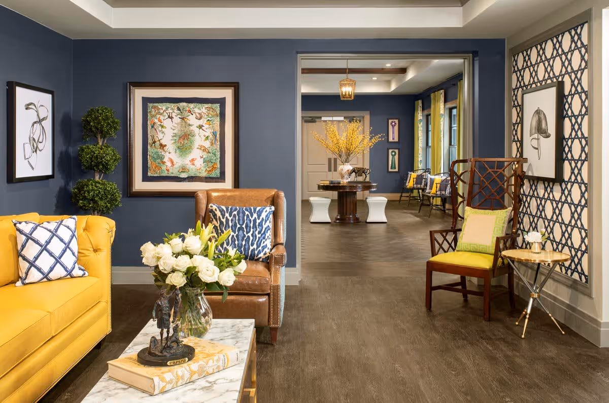 A stylish living room area with a yellow sofa and a brown leather armchair, both adorned with patterned cushions. A marble coffee table holds a vase of white flowers and a decorative sculpture. The walls are painted dark blue and decorated with framed artwork and a topiary plant. In the background, there is an open doorway leading to another room with a round table, yellow floral arrangement, and additional seating with yellow cushions.