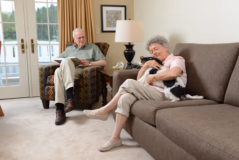 An elderly man sitting in an armchair reading a book and an elderly woman sitting on a sofa holding and cuddling a black and white cat in a well-lit living room with beige carpet, a table lamp, and a framed picture on the wall.