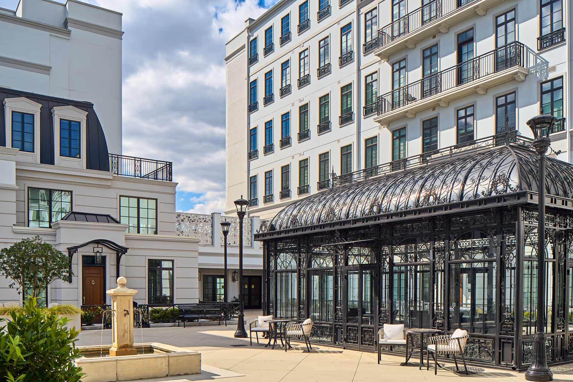 Outdoor courtyard area of a senior living facility featuring a decorative black wrought iron gazebo with glass panels, several chairs and tables, a stone water fountain, street lamps, and a multi-story white building with many windows in the background under a partly cloudy sky.