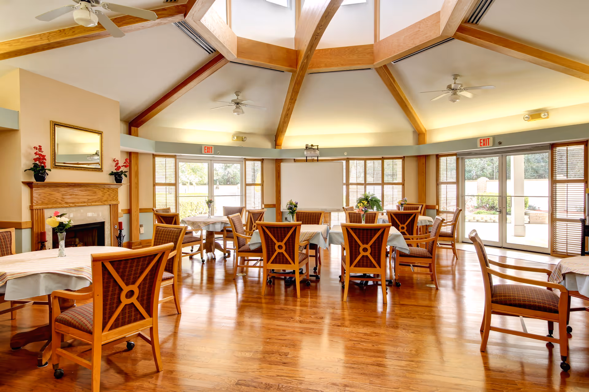 A bright and spacious common room with wooden floors and a high ceiling featuring exposed wooden beams. The room contains several round tables covered with tablecloths and surrounded by wooden chairs with patterned cushions. There are flower vases on the tables and a fireplace with a mirror above it on the left wall. Large windows and glass doors allow natural light to fill the room, and ceiling fans are mounted on the ceiling.