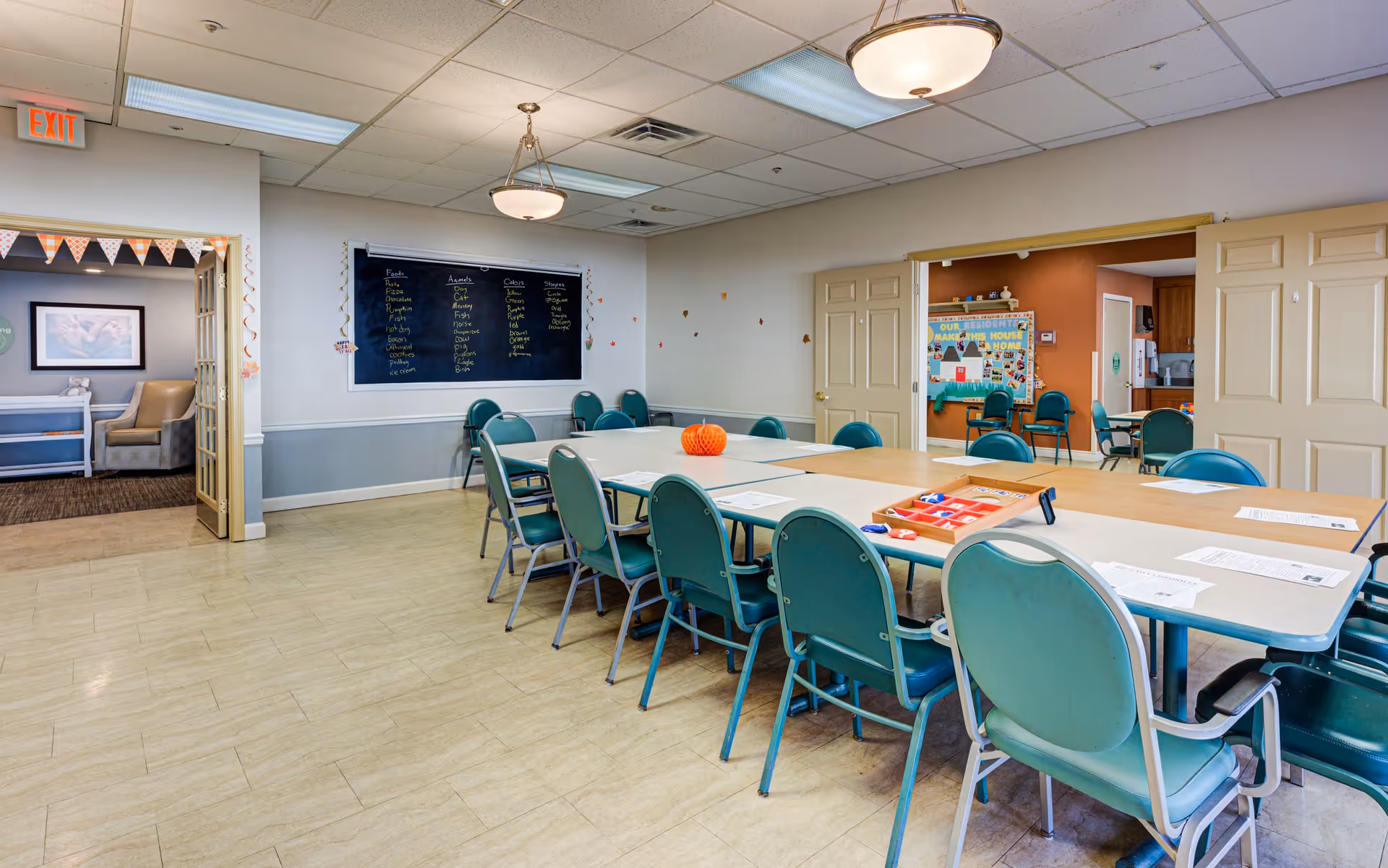 A well-lit activity or meeting room with a large rectangular table surrounded by teal chairs. On the table are papers, a small orange pumpkin decoration, and a game board. The walls are light-colored with a blackboard displaying names and activities. Double doors lead to another room with more chairs and colorful bulletin boards. An exit sign is visible above a doorway leading to a lounge area with armchairs and framed artwork.