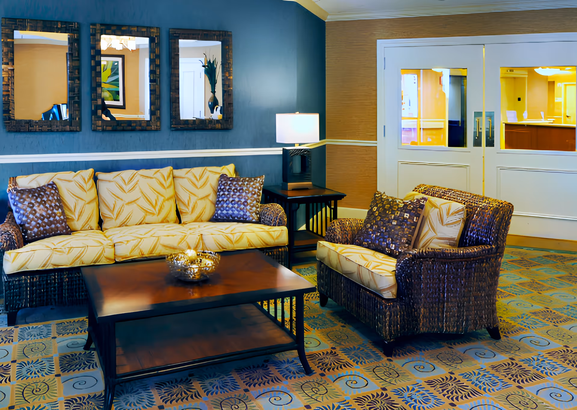 A furnished lounge with a wicker sofa and armchair, coffee table, lamp, and three decorative mirrors on a blue wall.