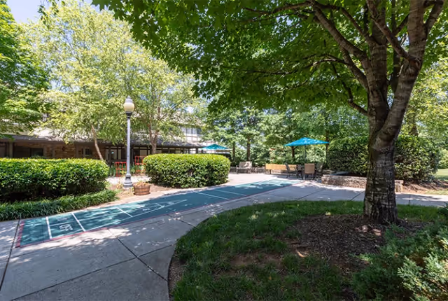 Outdoor patio area at Deane Hill Place Senior Living featuring a shuffleboard court, surrounded by green bushes and trees providing shade. There are benches and tables with blue umbrellas in the background, and a building partially visible behind the greenery.
