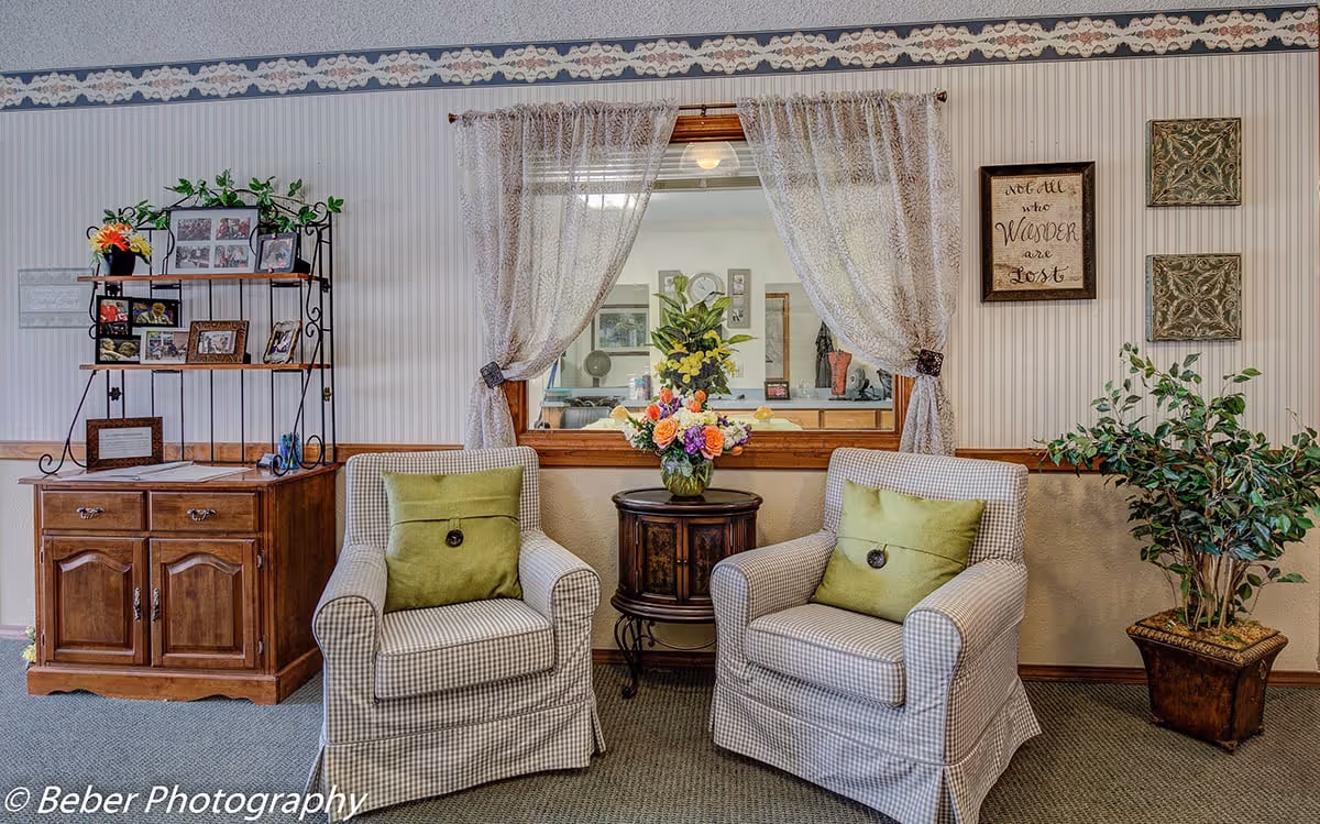 Two upholstered armchairs with green pillows flanking a small table with a flower arrangement in a decorated lounge area.