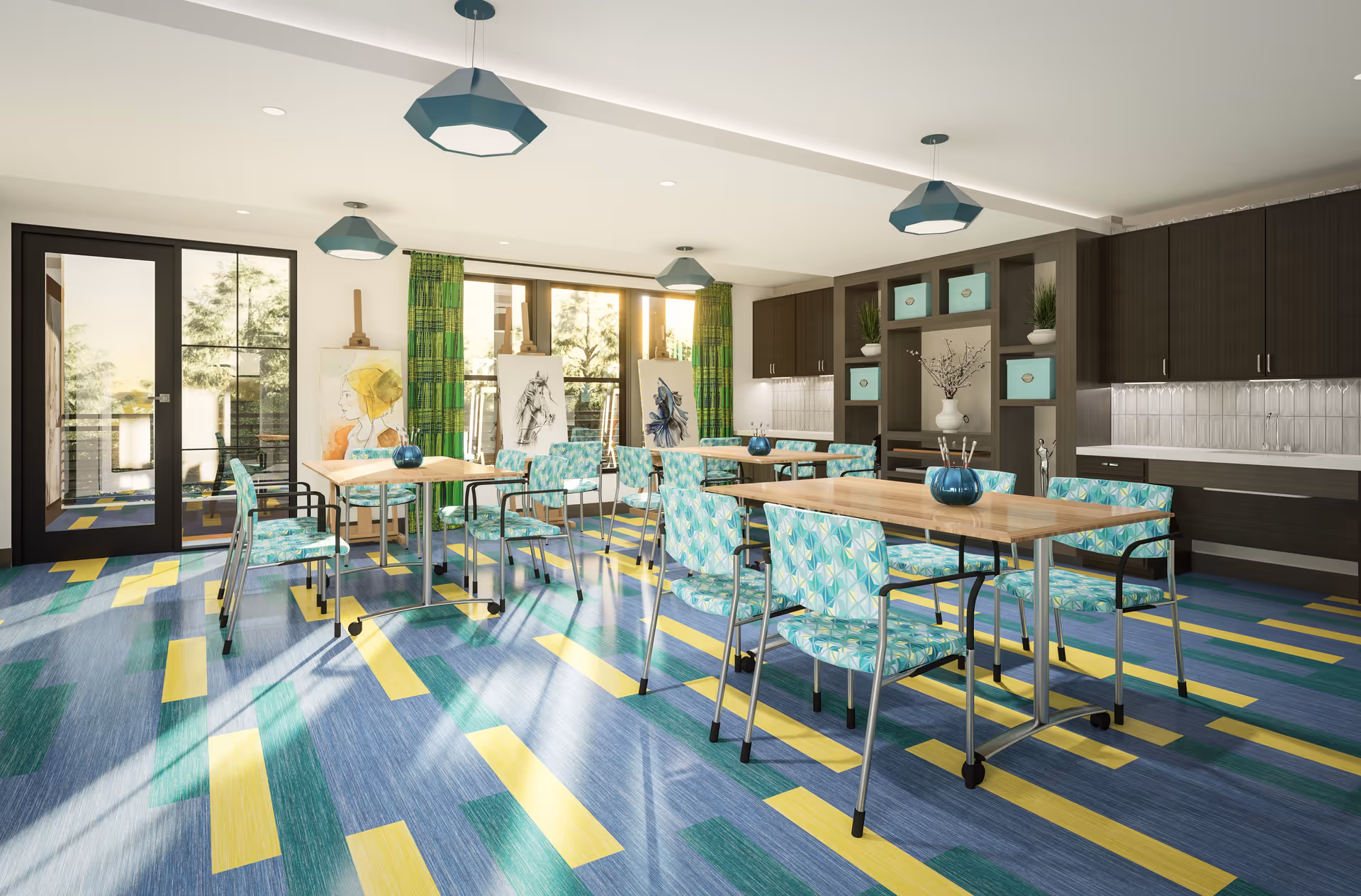 Bright and colorful activity room with multiple tables and chairs featuring blue and green patterned upholstery. The room has large windows with green curtains letting in natural light, three easels with artwork, modern pendant lights, and a kitchenette area with dark wood cabinets and a white countertop.