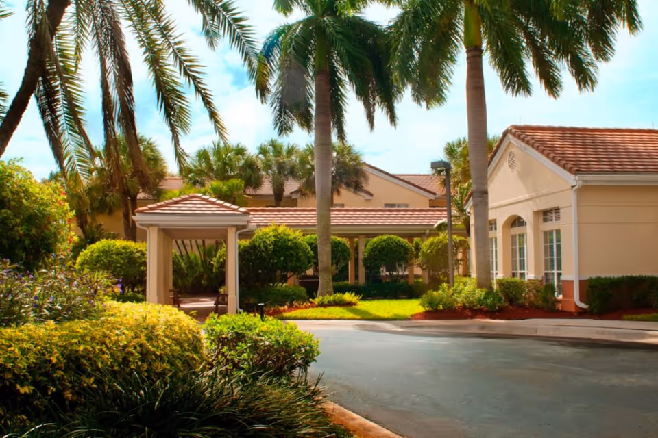 Outdoor view of Barrington Terrace facility featuring a driveway, lush green landscaping with bushes and palm trees, and beige buildings with red-tiled roofs under a partly cloudy sky.