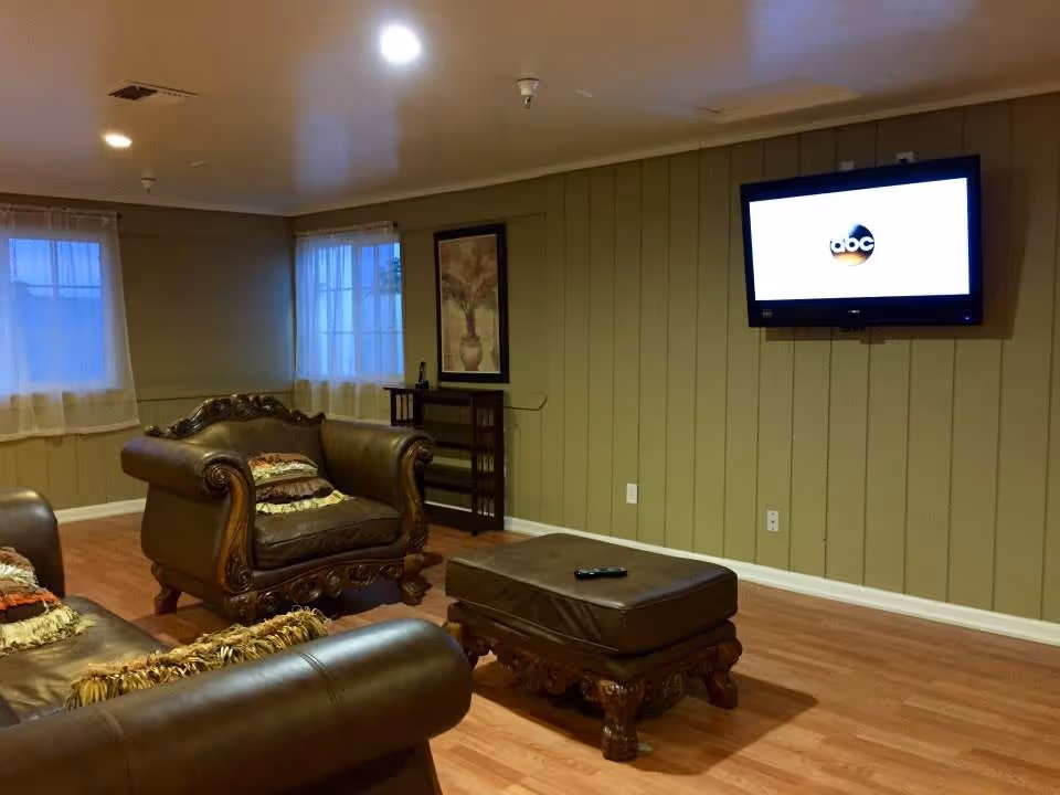 A cozy living room with wooden flooring, two brown leather armchairs with decorative pillows, a matching leather ottoman, a wall-mounted flat screen TV displaying the ABC logo, two windows with sheer curtains, a framed artwork on the wall, and a small wooden shelf unit.