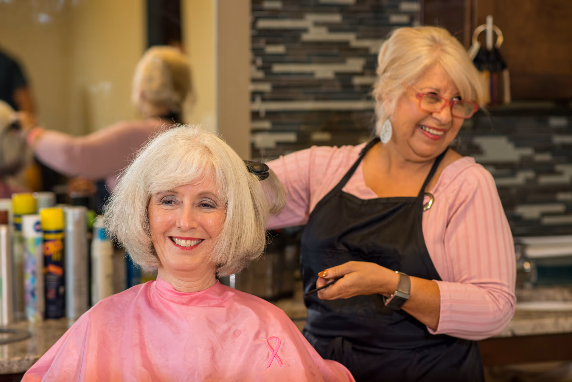 An elderly woman with white hair is sitting in a salon chair wearing a pink cape with a breast cancer awareness ribbon, smiling as a hairstylist with blonde hair and red glasses styles her hair. The background shows a countertop with hair products and a tiled backsplash.