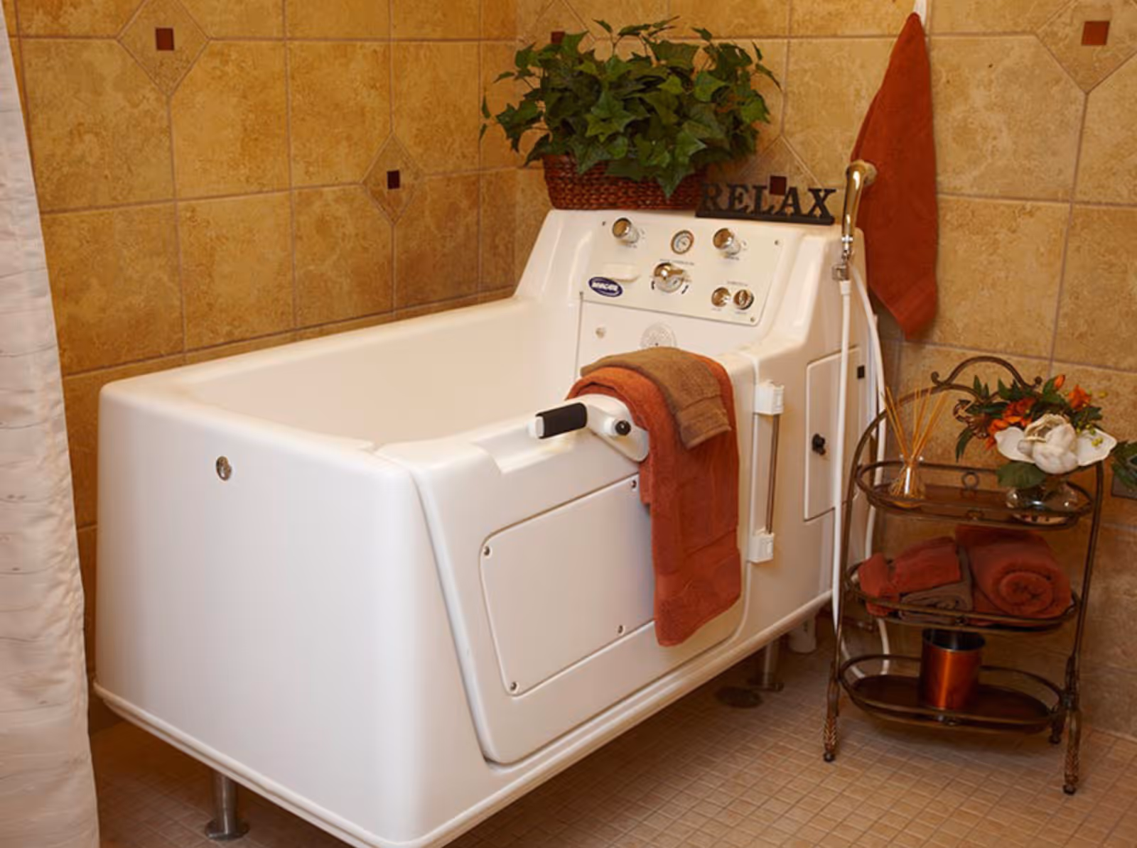 A walk-in bathtub with control knobs and a handle, surrounded by beige tiled walls. A red towel and a brown towel hang on the tub, and a green potted plant sits on the back edge. Next to the tub is a small metal cart with red towels, a flower arrangement, and reed diffusers. A red towel hangs on the wall behind the tub.