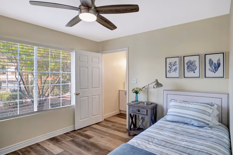 A bright bedroom with a single bed dressed in striped bedding, a nightstand with a lamp and a small vase of flowers, three framed botanical prints on the wall above the bed, a ceiling fan with wooden blades, and a large window with white blinds showing greenery outside. An open door leads to a small adjacent room with a sink.