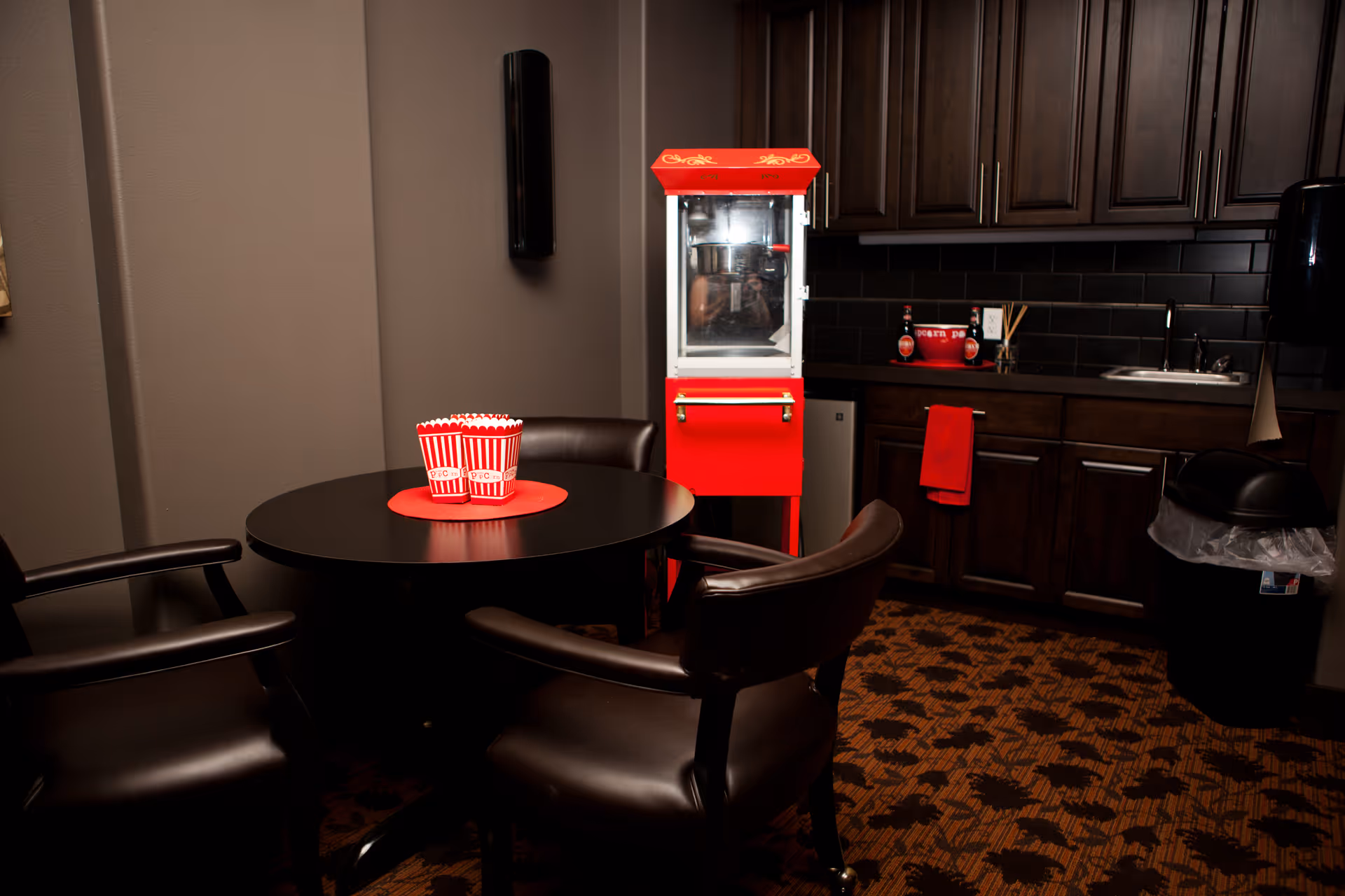 A small kitchenette area with dark wood cabinets, a sink, and a black trash can. In front of the cabinets is a red popcorn machine. A round black table with three black leather chairs surrounds it, with three red and white striped popcorn containers on a red placemat on the table. The floor has a patterned carpet with dark and light brown colors.