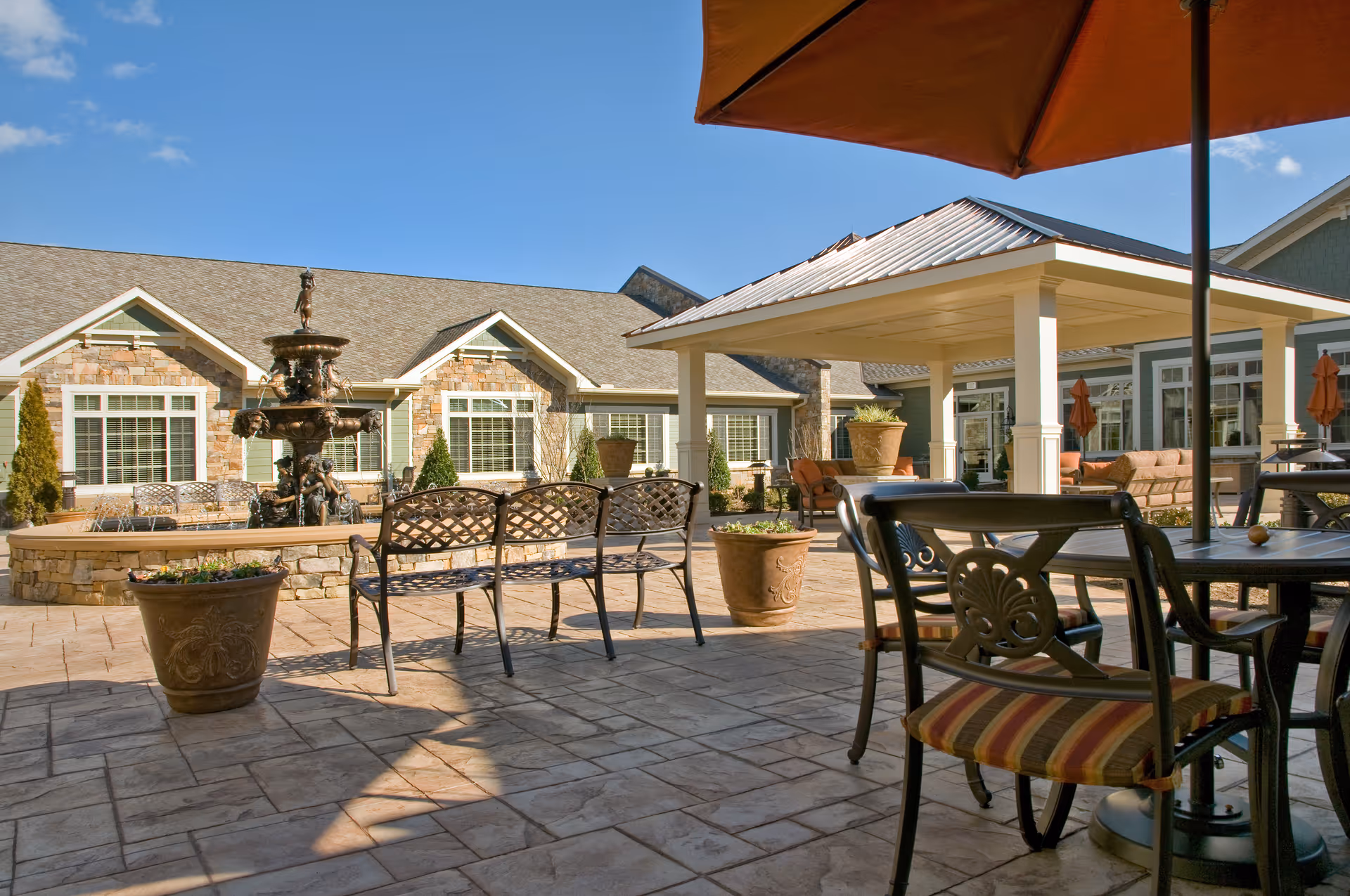 Outdoor patio area at a senior living facility with metal benches, tables with chairs, large potted plants, a multi-tiered fountain, and a covered seating area under a clear blue sky.