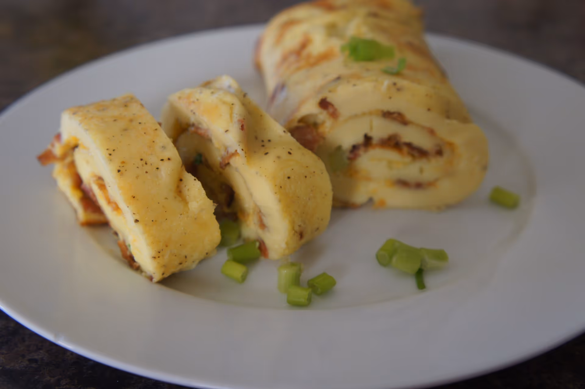 A white plate with a rolled omelette sliced into pieces, garnished with chopped green onions on a dark surface.