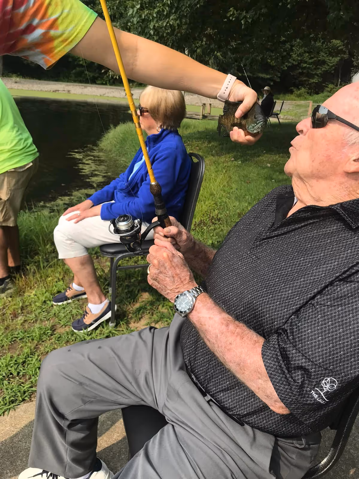 An elderly man sitting on a chair near a pond holding a fishing rod, reacting with surprise as a person in a tie-dye shirt holds a small fish close to his face. Another elderly woman in a blue jacket and white pants sits nearby, also by the pond.