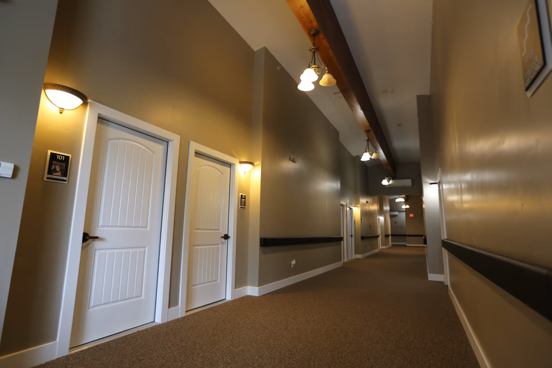 A long indoor hallway with beige walls and carpeted floor, featuring multiple white doors with room numbers and small wall-mounted lights above each door. The ceiling has exposed wooden beams with hanging light fixtures.
