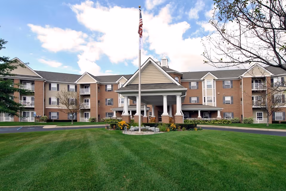 Front exterior view of Elmhaven Manor, a three-story brick building with multiple windows and balconies. There is a covered entrance with white columns and a flagpole with an American flag in the center of a well-maintained green lawn. Trees and shrubs surround the building under a partly cloudy sky.