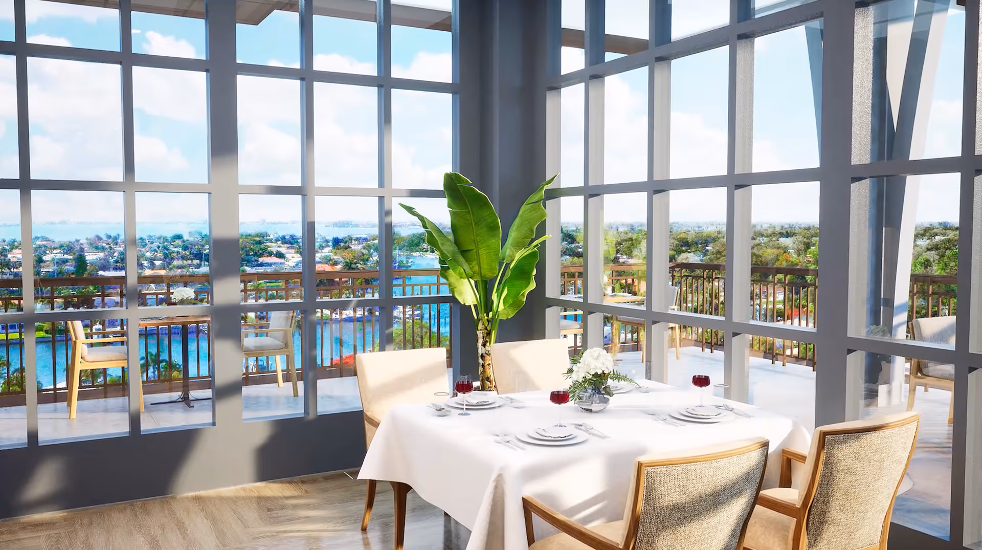 A bright dining area with a table set for four, featuring white tablecloth, plates, cutlery, and glasses with red liquid. The room has large floor-to-ceiling windows offering a scenic view of a waterfront and surrounding neighborhood. There is a large green plant near the window and additional seating visible outside on a balcony.