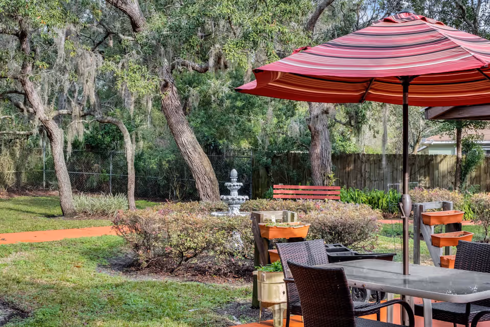 Outdoor courtyard with a striped red umbrella over a table and chairs, nearby fountain, bench, planters and trees.