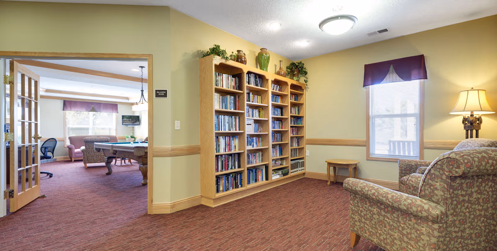 Interior view of a senior living facility common area with a bookshelf filled with books, a patterned armchair, a small wooden table, a lamp, and a window with blinds and a purple valance. Through an open door with glass panels, a billiards room is visible with a pool table, chairs, a sofa, and a wall-mounted TV.