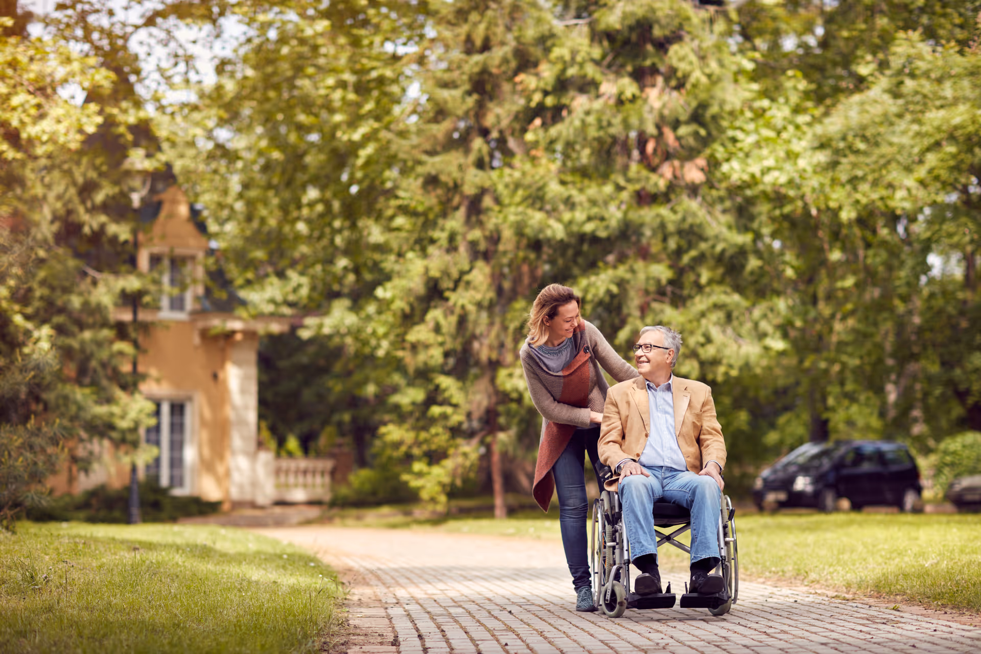 A woman is pushing an elderly man in a wheelchair along a paved path in a green, leafy outdoor setting with trees and a building in the background. Both are smiling and appear to be enjoying the pleasant weather.