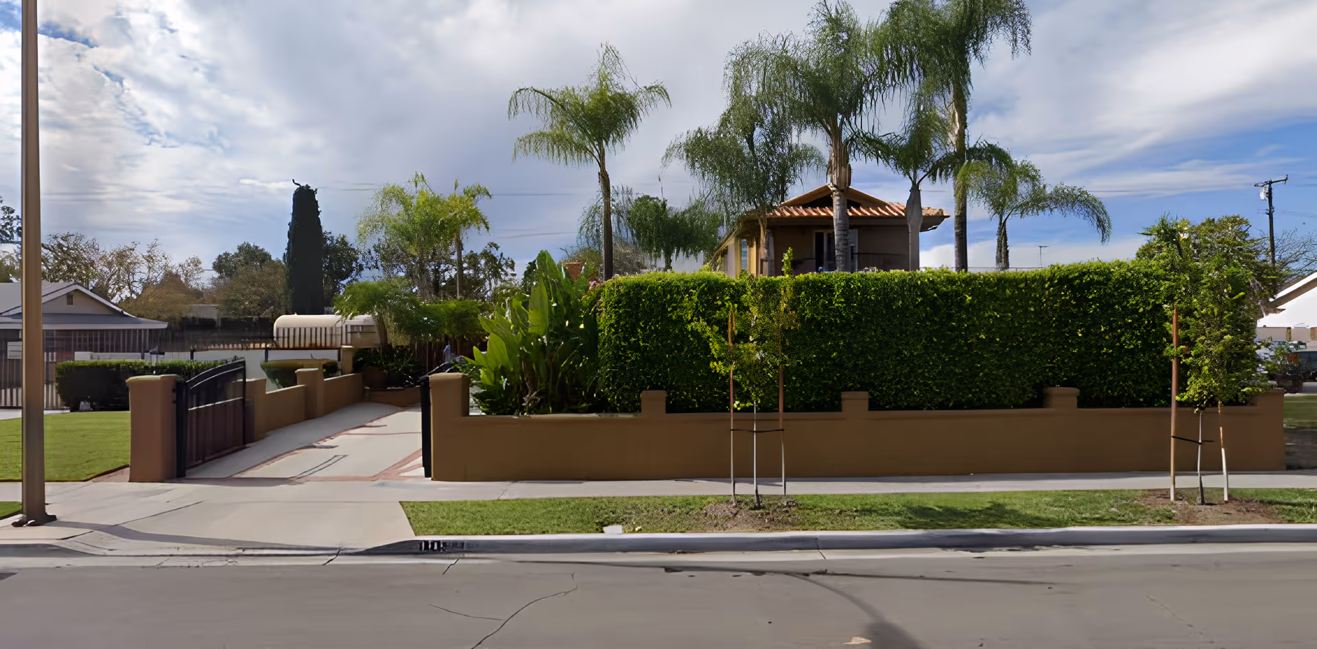 Exterior view of a senior care facility with a tall green hedge, palm trees, and a driveway gate. The building has a tiled roof and is partially obscured by the hedge. The sky is partly cloudy.