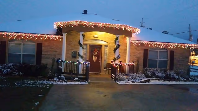 Single-story building entrance decorated with holiday lights, a wreath on the door, and light snow on the ground at dusk.