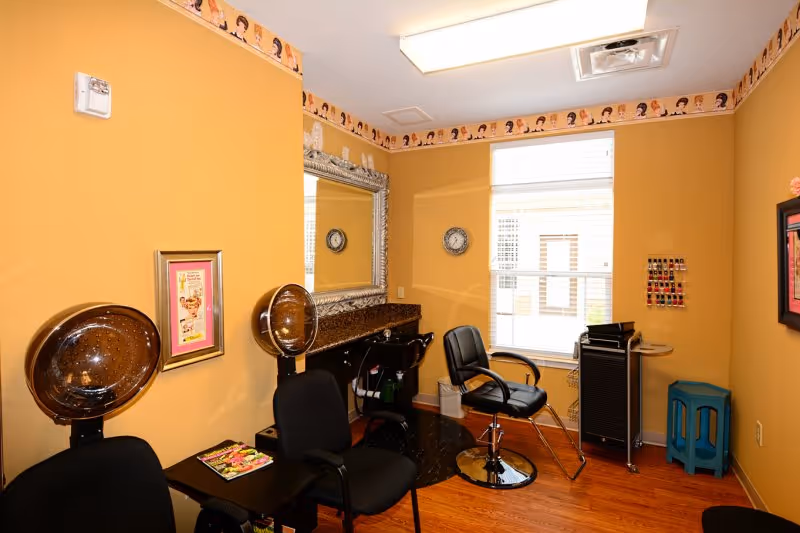 Interior of a small salon room with mustard yellow walls, two black salon chairs with hair dryers, a large ornate mirror above a granite countertop with a sink, a black rolling cart, a small blue stool, a window with blinds, and framed pictures on the walls.
