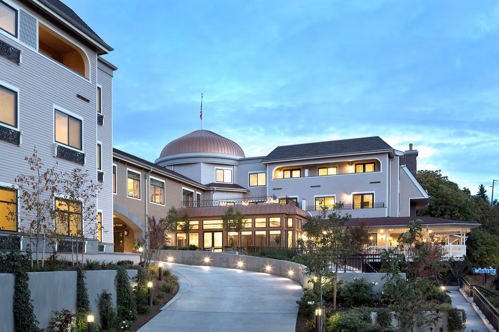 Exterior view of Aegis Living Queen Anne Rodgers Park building at dusk, showing a curved driveway leading to the entrance with well-lit windows and landscaped greenery along the path.