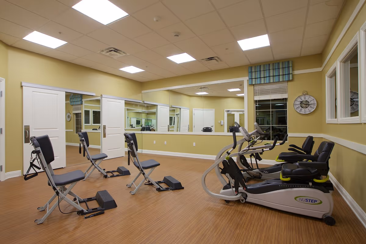 A fitness room with exercise equipment including three recumbent exercise bikes and three folding exercise chairs. The room has light yellow walls, a large mirror on one wall, a window with a blue and green striped valance, and a clock on the wall. The floor is wood-patterned, and the ceiling has recessed lighting.