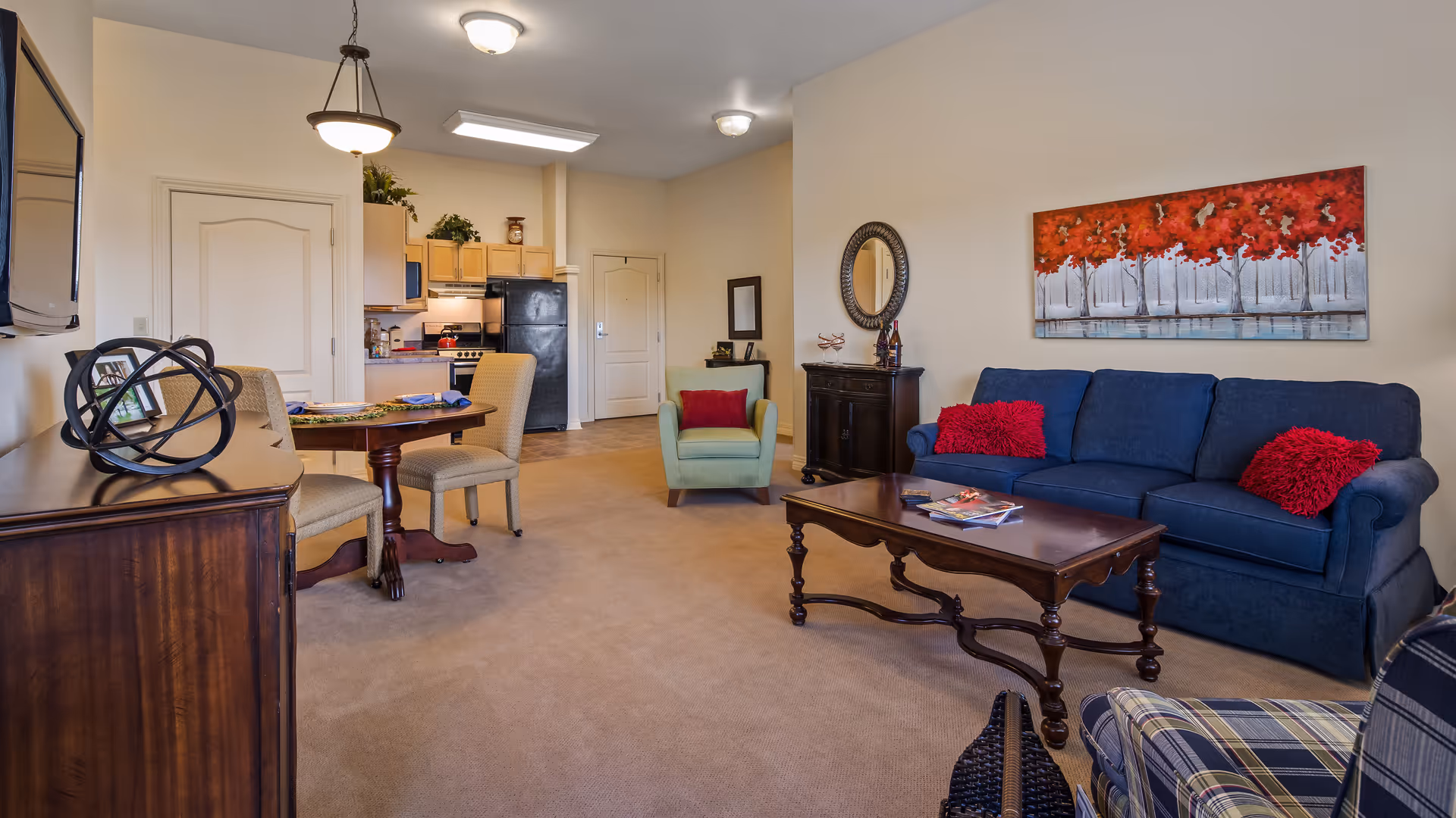 A cozy living room and kitchen area in an apartment at The Regent. The living room features a blue sofa with red pillows, a wooden coffee table with magazines, a green armchair, and a plaid armchair. A colorful painting of red trees hangs on the wall above the sofa. The kitchen area has light wood cabinets, a black refrigerator, and a stove. A small dining table with two beige chairs is positioned near the kitchen. The room is carpeted and well-lit with ceiling lights.