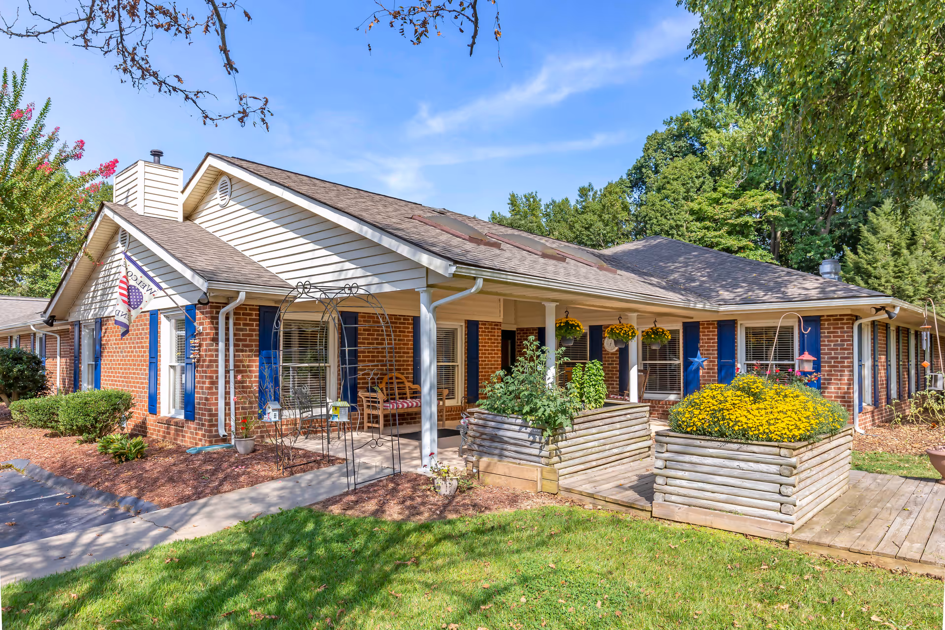 Exterior view of a single-story brick building with white siding and blue shutters, featuring a covered porch with hanging flower pots, a wooden bench, and raised garden beds with yellow flowers and greenery. The building is surrounded by a well-maintained lawn, trees, and a clear blue sky.