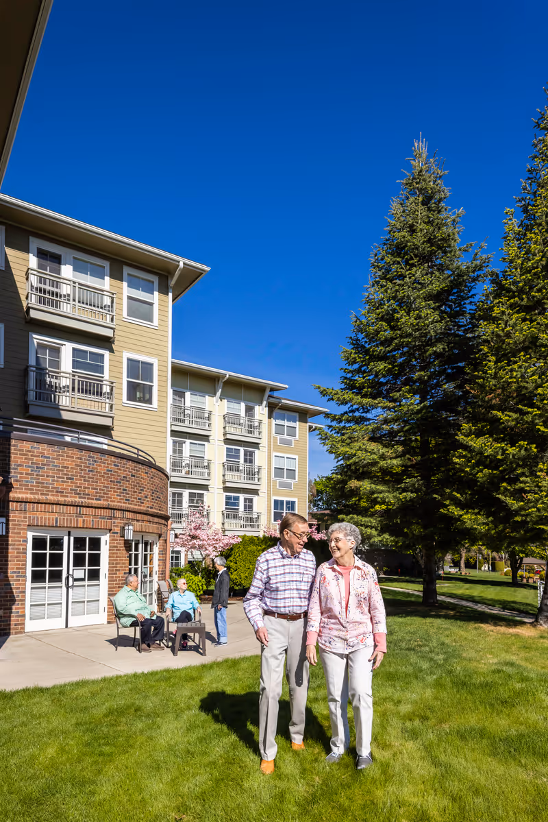 Two elderly people walking on a grassy lawn in front of a multi-story senior living building while other residents sit on a patio under tall trees.