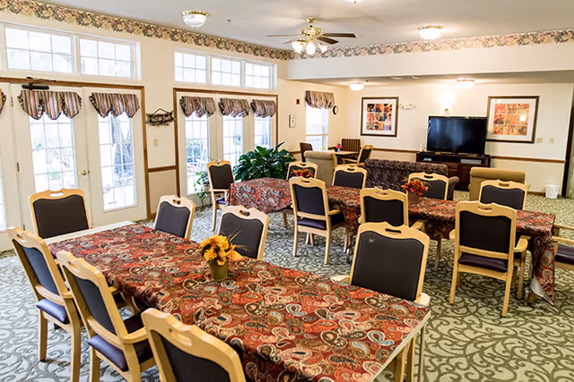 A bright and spacious common area in a senior living facility with multiple tables covered in patterned red tablecloths, surrounded by chairs. The room has large windows and glass doors letting in natural light, floral wallpaper border near the ceiling, ceiling fans, and a television on a stand against the far wall. There are framed pictures on the walls and a green patterned carpet.