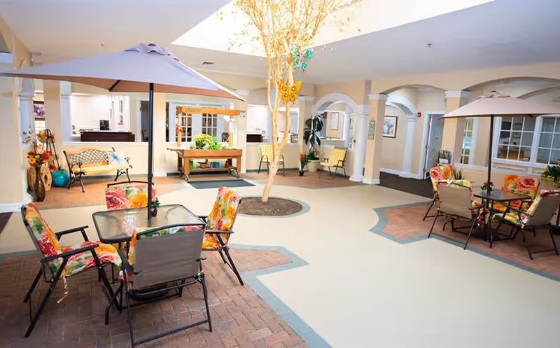 Indoor common area with seating arrangements including tables with umbrellas and chairs with floral cushions, a tree planted in the center, benches along the walls, and decorative elements such as plants and wall art.