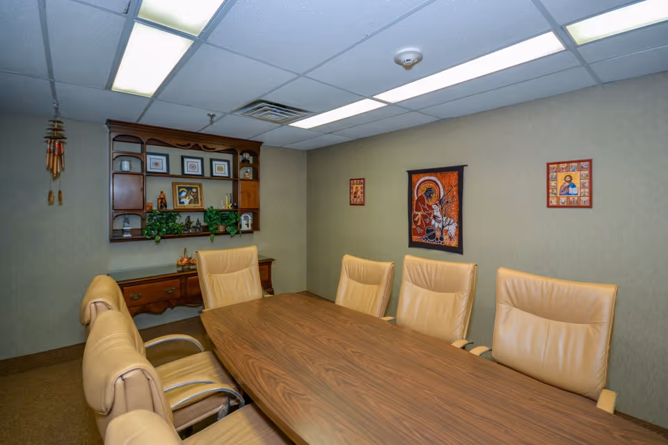 A conference room with a long wooden table surrounded by six beige leather chairs. The walls are decorated with framed artwork and a wooden shelf holding small decorative items and plants. The ceiling has fluorescent lighting panels.