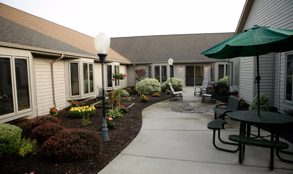 Outdoor courtyard area of a senior living facility with a curved concrete walkway, garden beds with various shrubs and flowers, several black metal chairs and tables, and green umbrellas providing shade. The building has beige siding and multiple windows facing the courtyard.