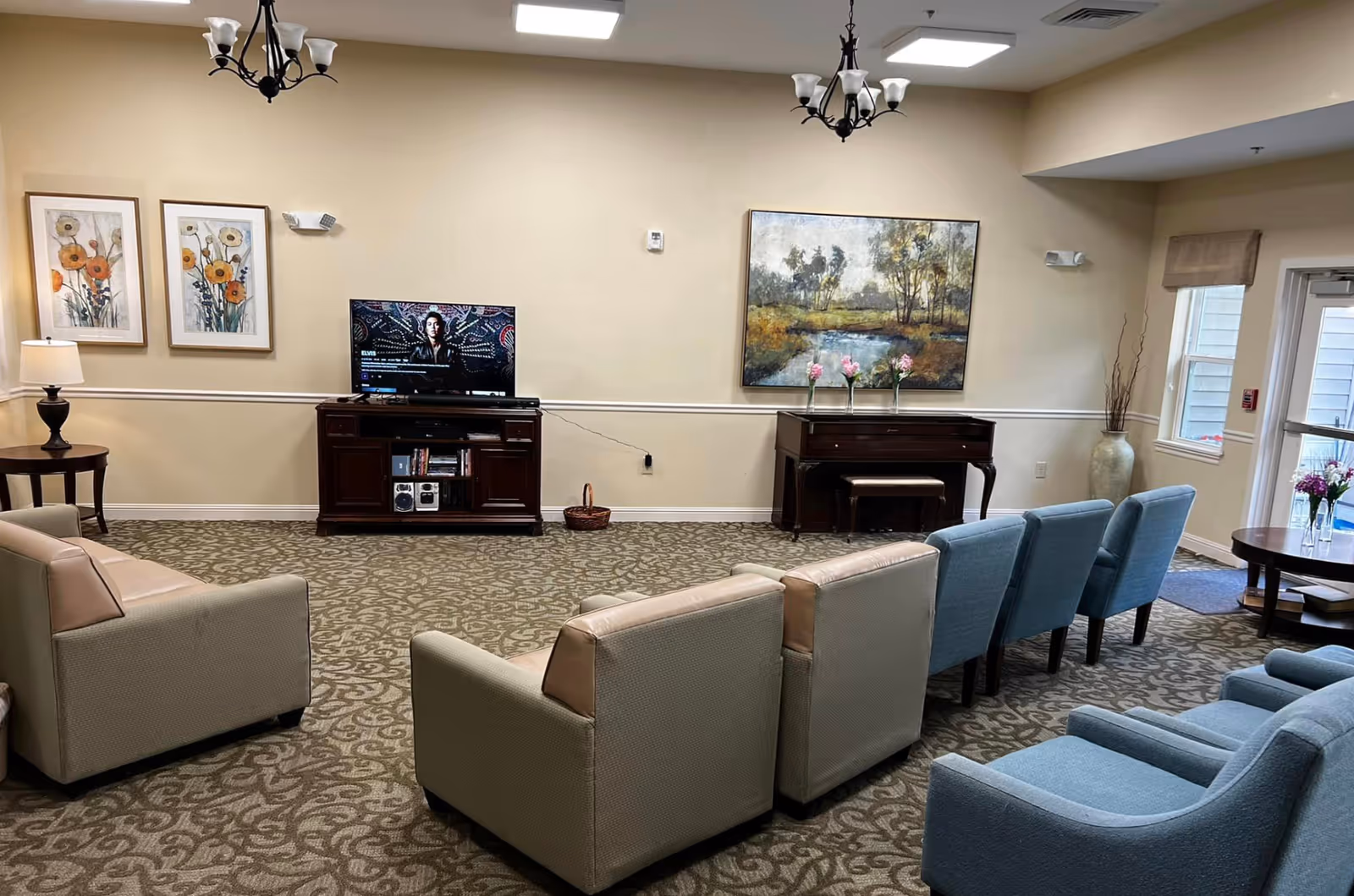 A senior living facility common area with beige and blue armchairs arranged facing a TV on a wooden stand. The room has patterned carpet, two chandeliers, framed floral and landscape paintings on the walls, a piano with flowers on top, a side table with a lamp, and windows letting in natural light.