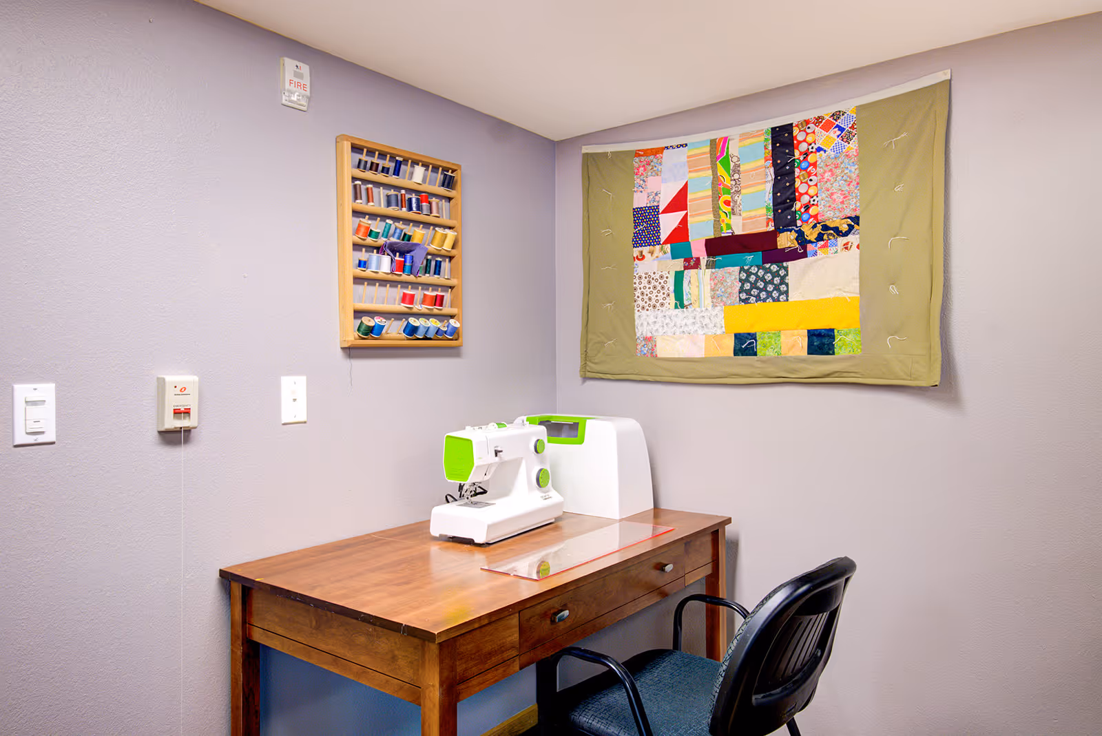 A small sewing area with a wooden table holding a white and green sewing machine. Above the table on the wall is a wooden rack filled with colorful spools of thread. A patchwork quilt hangs on the adjacent wall. A black chair is positioned in front of the table.