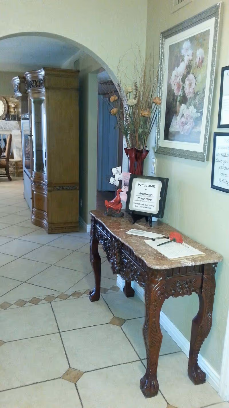 An interior hallway area with a decorative wooden table featuring intricate carvings and a marble top. On the table, there is a red vase with dried flowers, a small red bird figurine, a welcome sign for Greenway Home Care, and some papers with a pen. The wall behind the table has framed artwork and a notice. In the background, there is an archway leading to another room with wooden furniture and a fireplace.