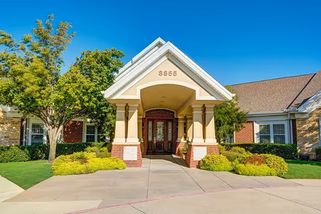 Front entrance of a building with a covered porch supported by columns, surrounded by green shrubs and trees under a clear blue sky. The building has a brick and stone exterior with the number 8555 displayed above the entrance.